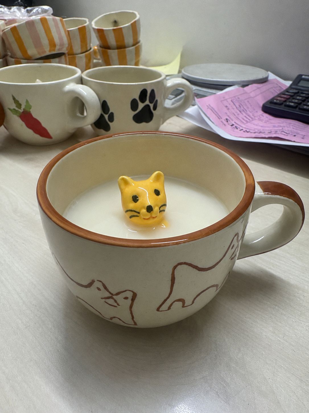 Cat-shaped toy in a mug of milk on a table with various items in the background.