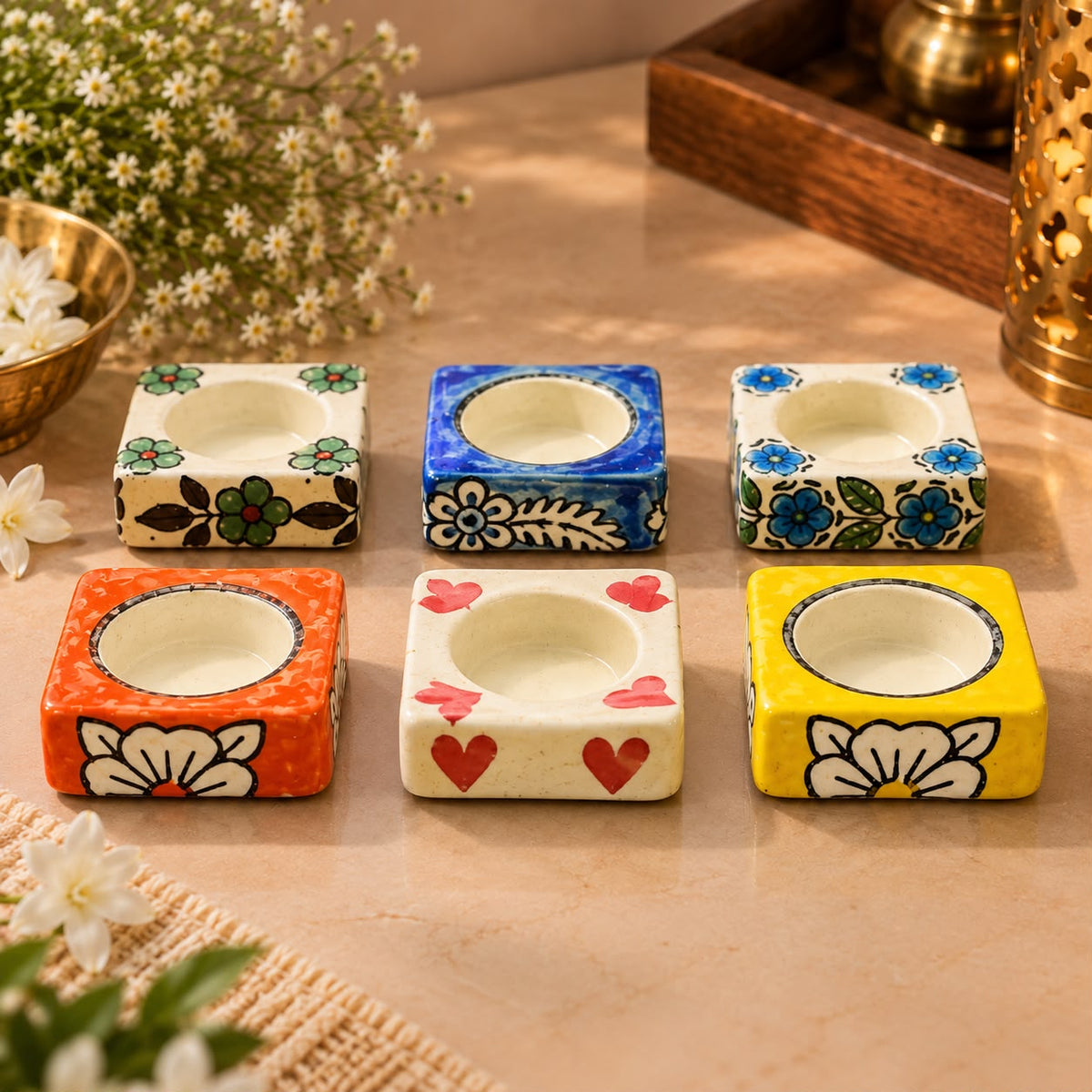 Six small square ceramic candle holders with floral and heart patterns in orange, blue, green, white, and yellow, arranged on a beige tabletop with decorative flowers and a brass lantern in the background.