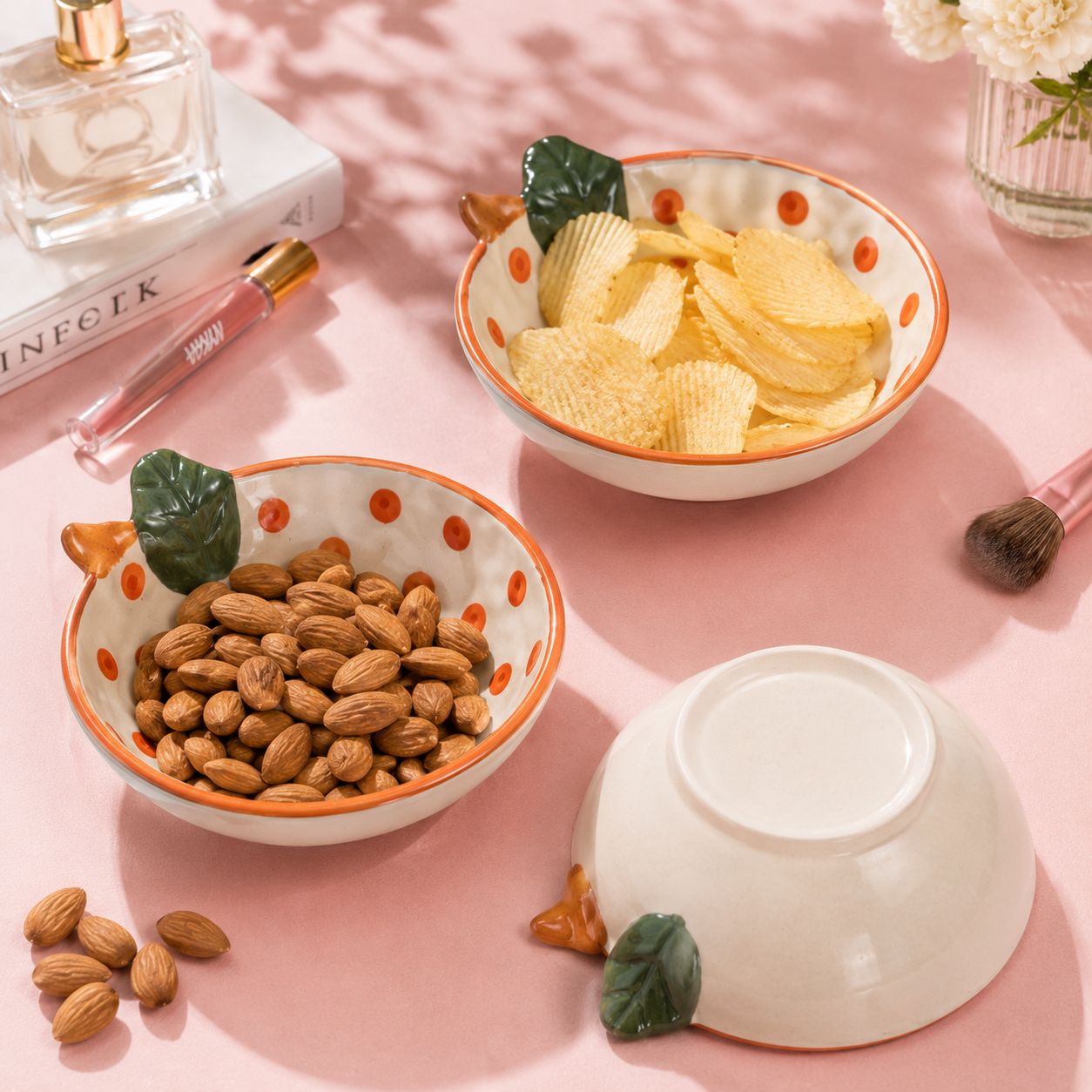 Two ceramic bowls with decorative handles, one containing almonds and the other potato chips, on a pink surface with a perfume bottle and makeup brush.