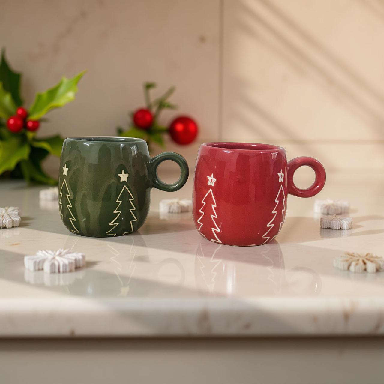 Two ceramic mugs, one green and one red, with Christmas tree designs on a marble surface.