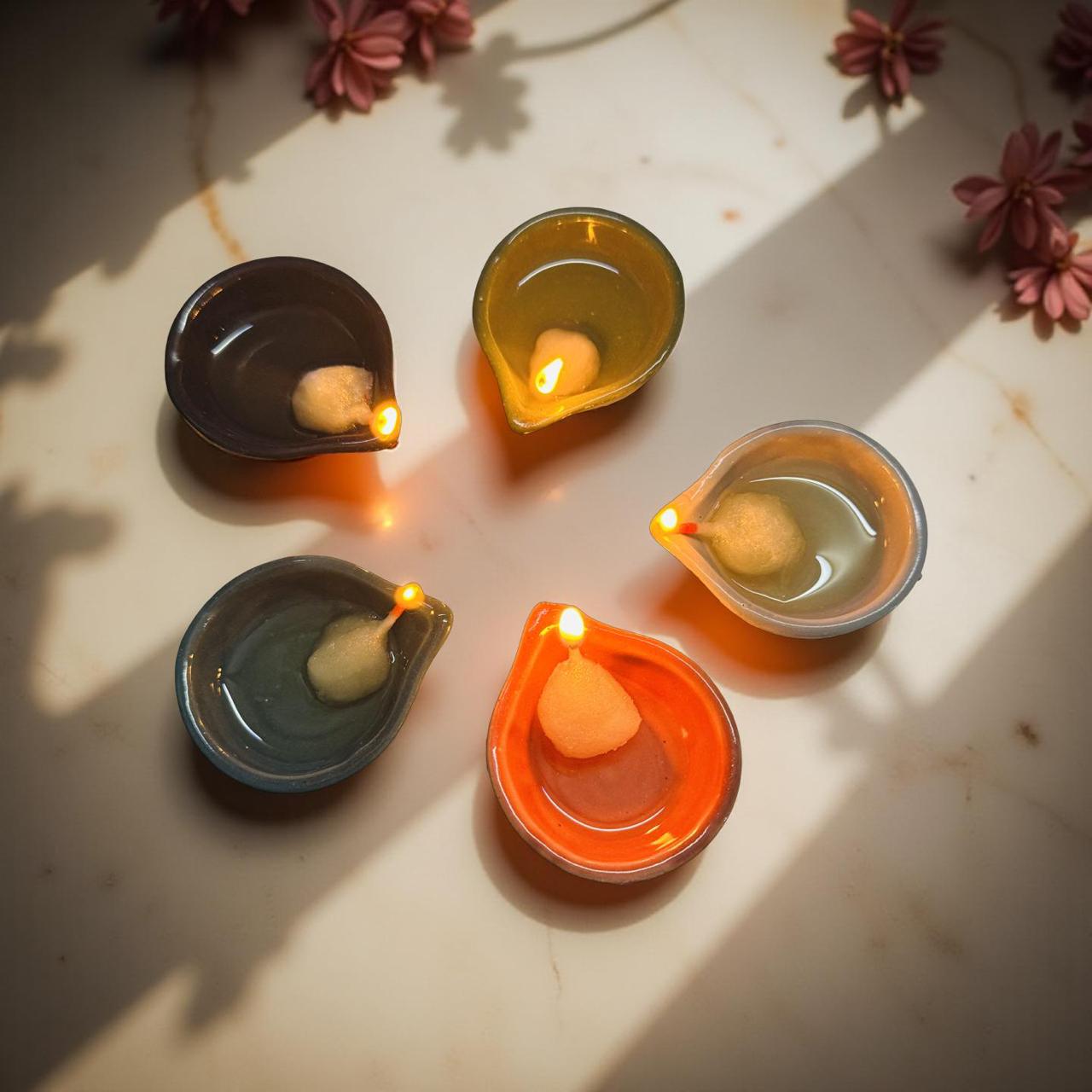 Five small ceramic lamps with lit candles on a light surface with flowers in the background