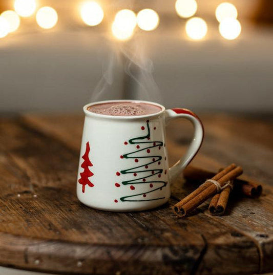 White mug with red and green designs on a wooden table with cinnamon sticks, against a blurred background of lights.