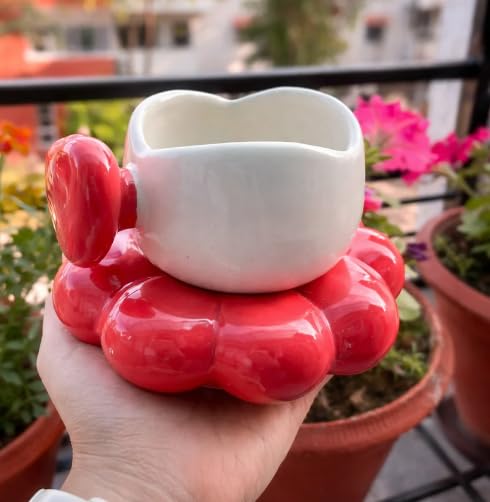 A hand holding a glossy ceramic cup and saucer set. The cup is white with a heart-shaped rim, resting on a red saucer shaped like a flower or bubble cluster.