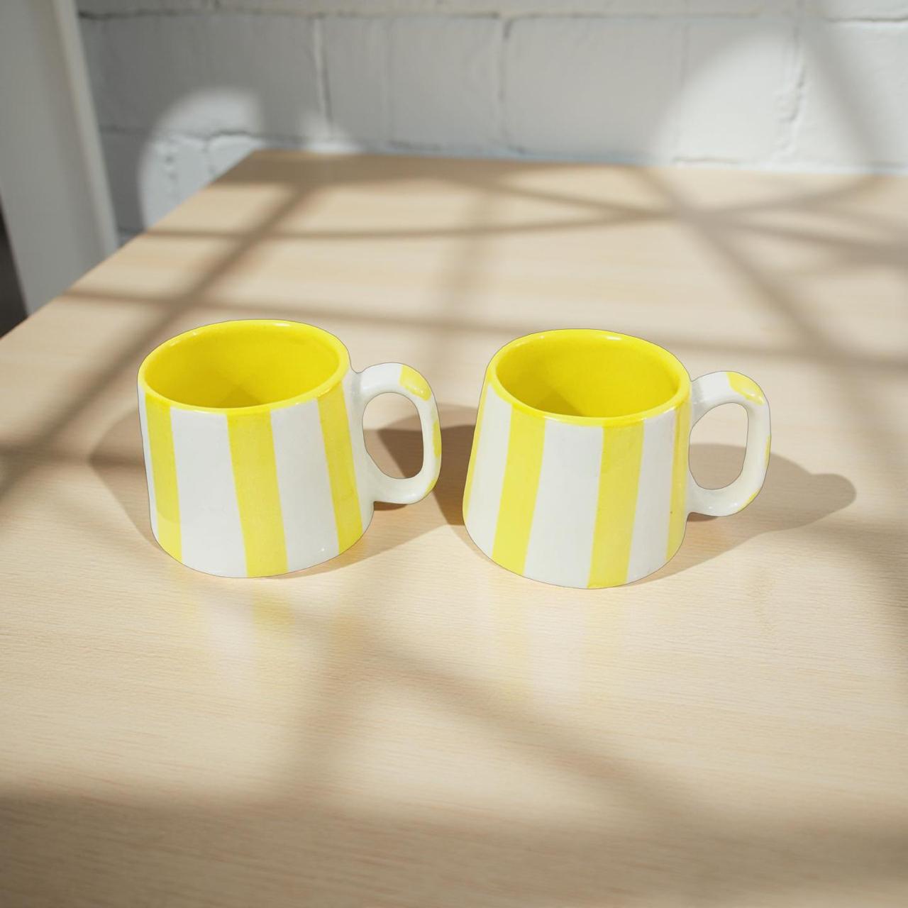 Two yellow and white striped mugs on a wooden table with sunlight casting shadows.
