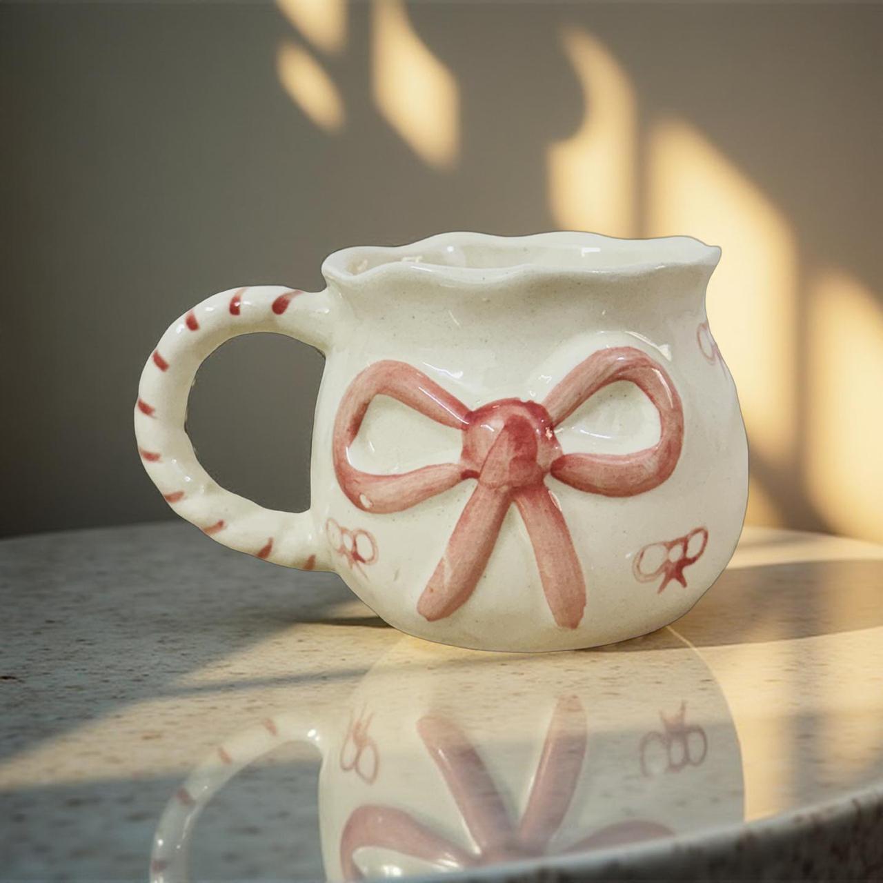 White ceramic mug with a pink bow design on a reflective surface.