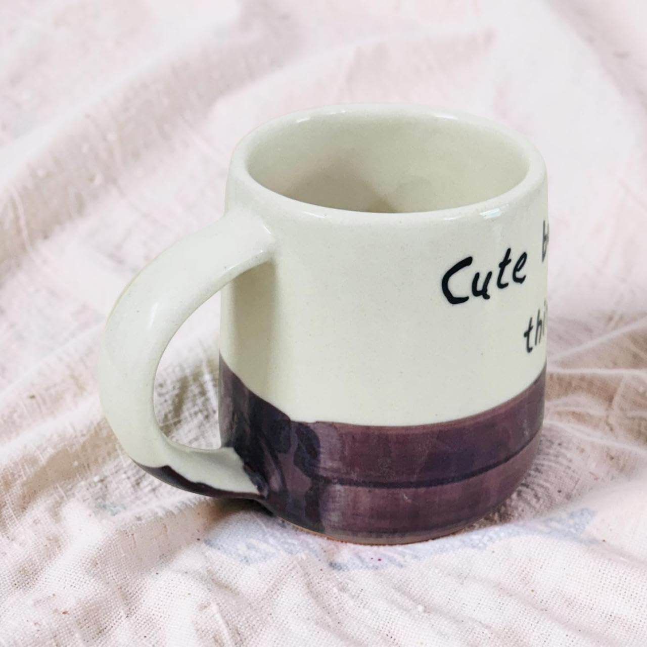 A ceramic mug with a cream-colored upper body, a dark purple painted lower band, and black printed text on the side, resting on a textured light pink fabric surface.