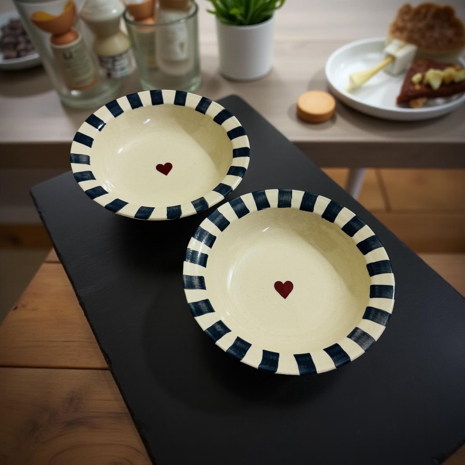 Two cream ceramic bowls with navy stripes and red heart center, placed on wooden table