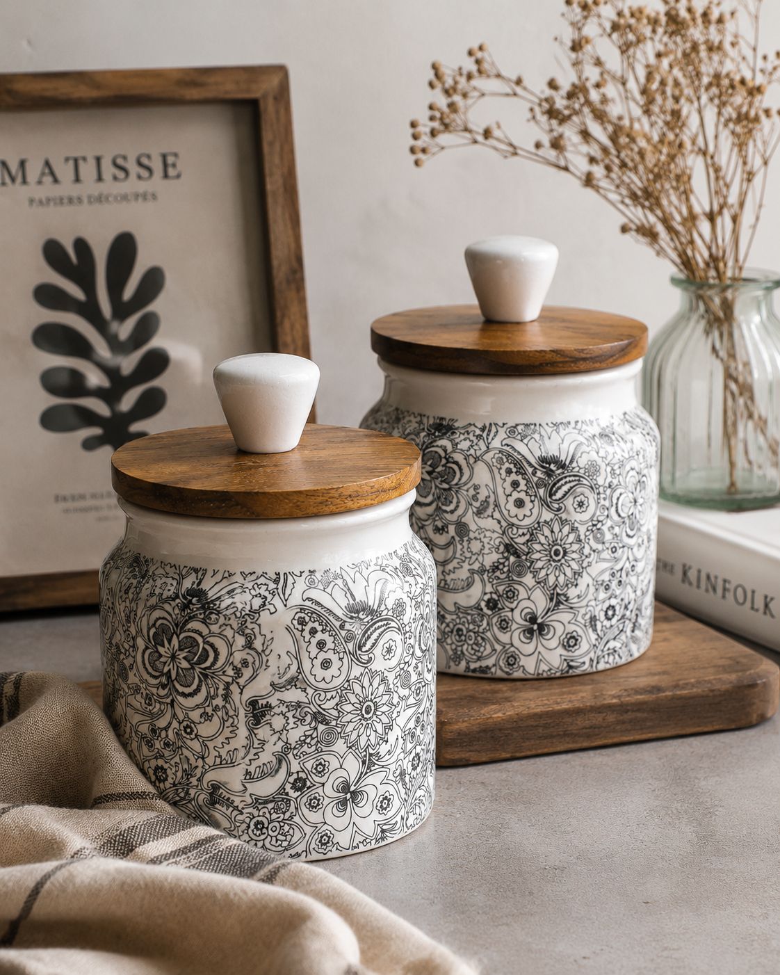 Two decorative jars with wooden lids on a surface with a framed picture and dried plants in the background.