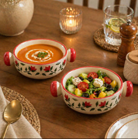Two decorative ceramic bowls with red handles sit on a wooden dining table. One bowl contains orange soup with a swirl of cream, and the other contains a colorful salad.