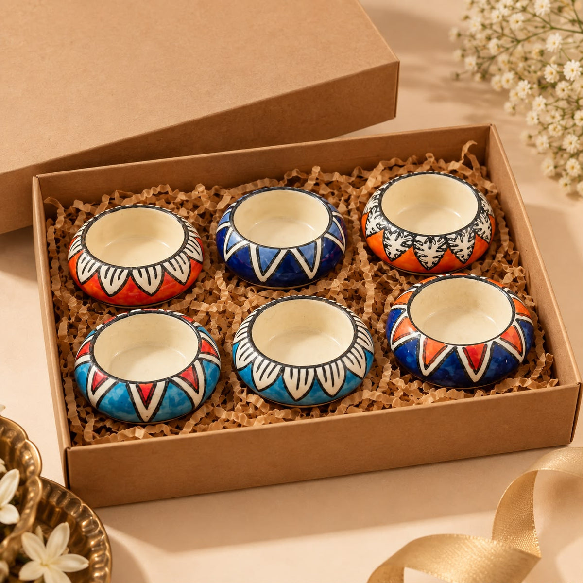 Six small hand-painted ceramic cups with colorful geometric patterns are arranged in a brown gift box filled with shredded paper. The cups feature blue, red, orange, and white designs, with cream-colored interiors.