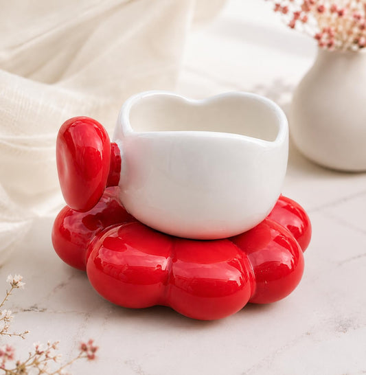A glossy white ceramic heart-shaped cup with a red handle, resting on a matching red flower-shaped saucer, set against a soft beige fabric background with a blurred vase of flowers.