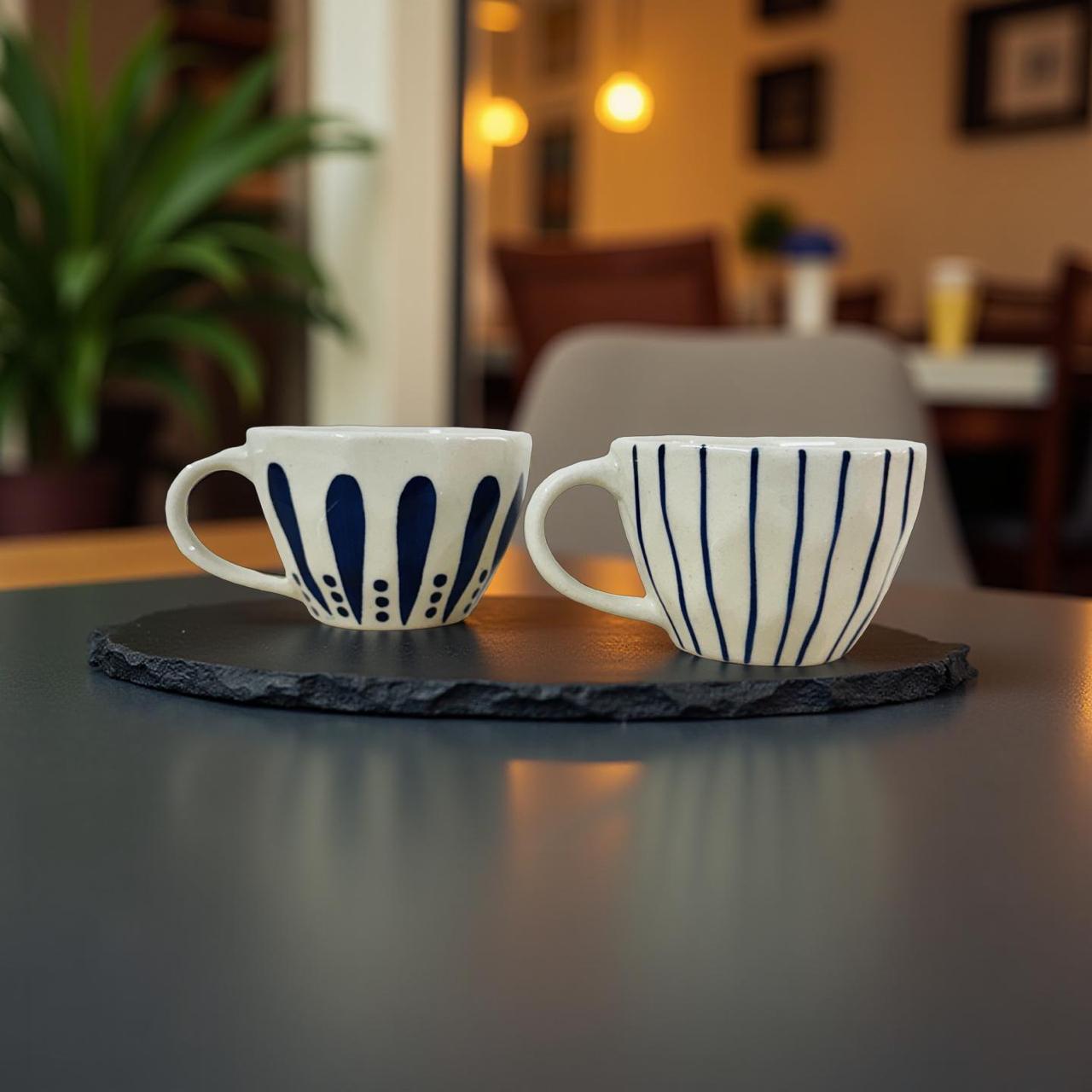 Two ceramic cups with blue patterns on a dark slate coaster in a blurred indoor setting.
