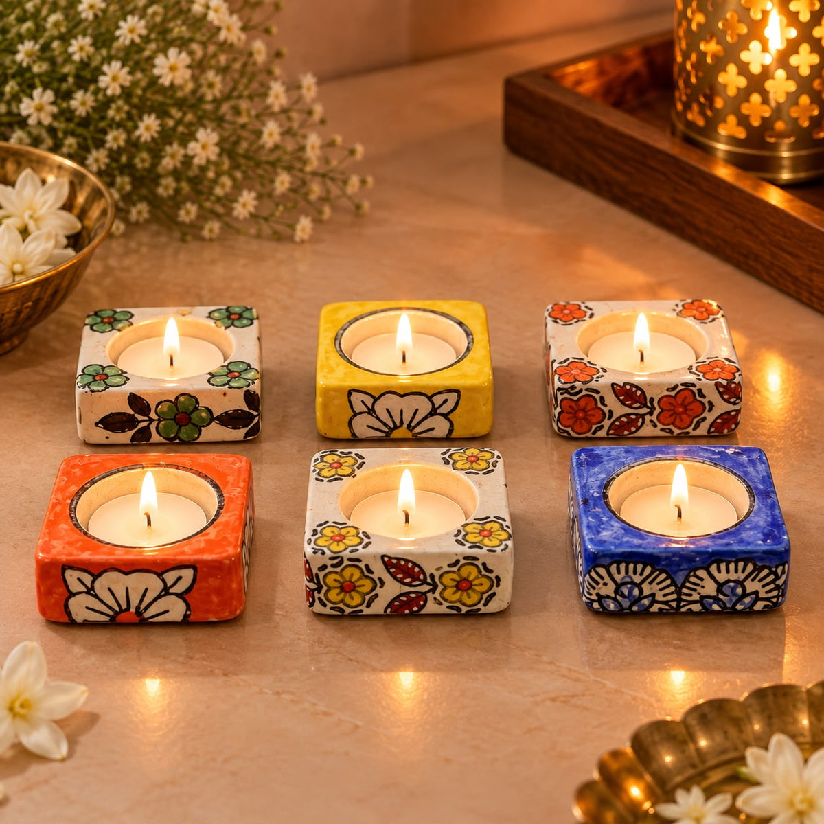 Six small square ceramic tea light candle holders with colorful floral patterns are arranged on a tabletop, each holding a lit tealight candle. The holders come in orange, yellow, and blue, with white, yellow, red, and green floral designs.