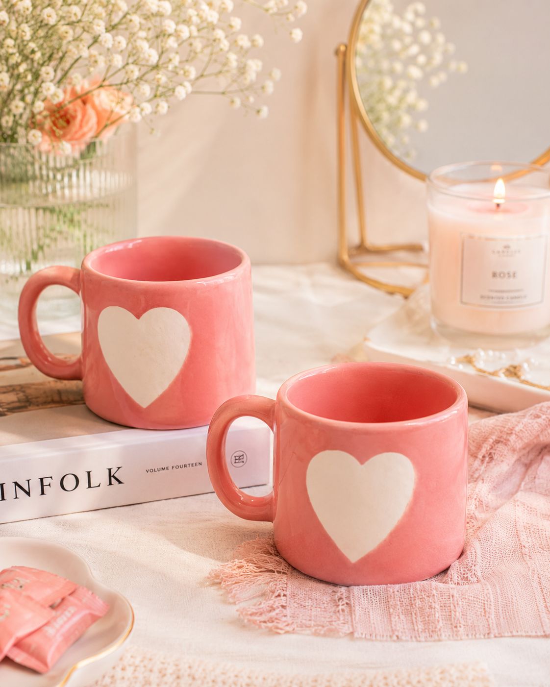 Two pink ceramic mugs with a white heart design on the front, styled on a table with a book, dried flowers, a candle, and a mirror in a soft, warm setting.