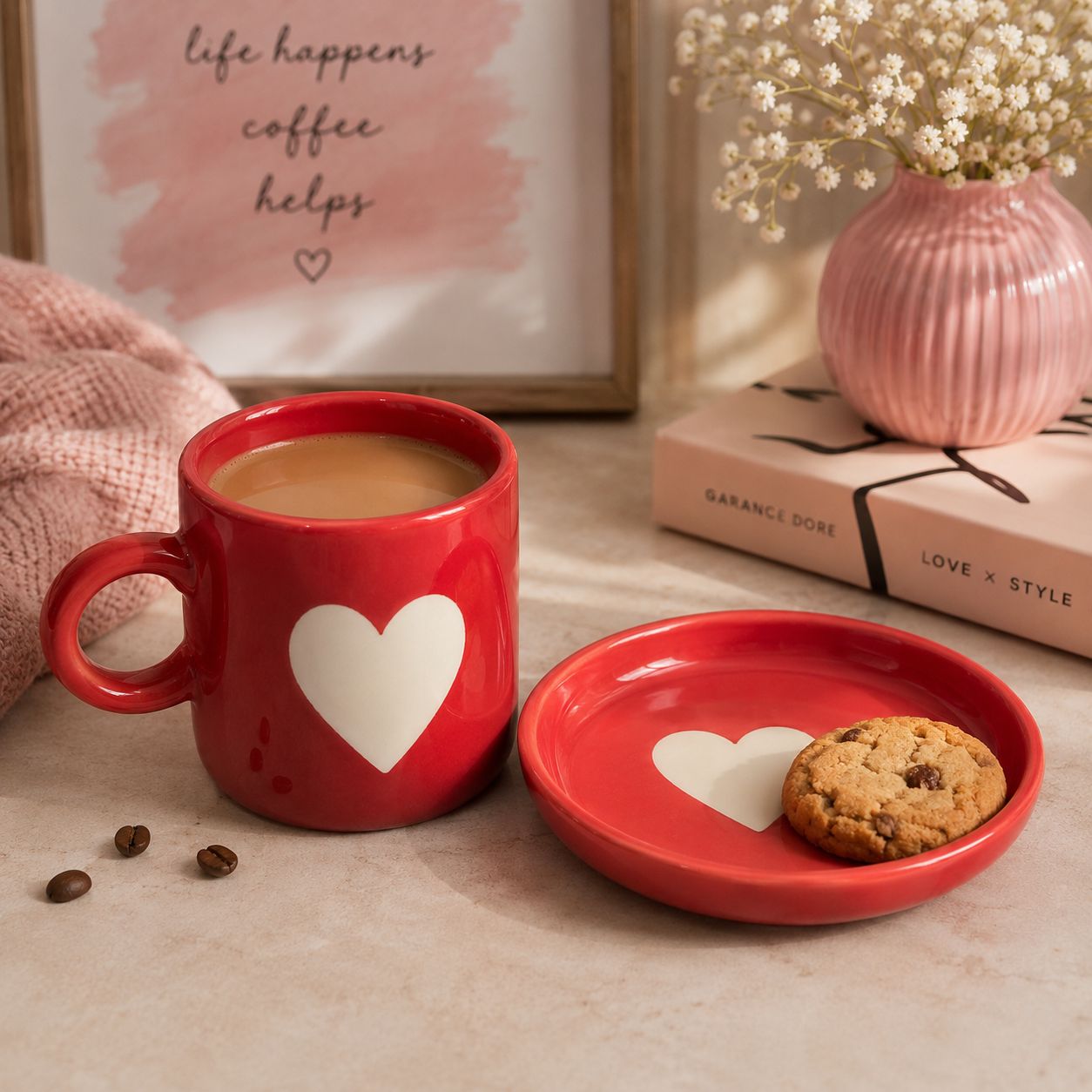 A red ceramic mug filled with coffee and a matching small red saucer with a white heart, styled on a tabletop with a cookie, books, a pink vase, and a framed print in the background.