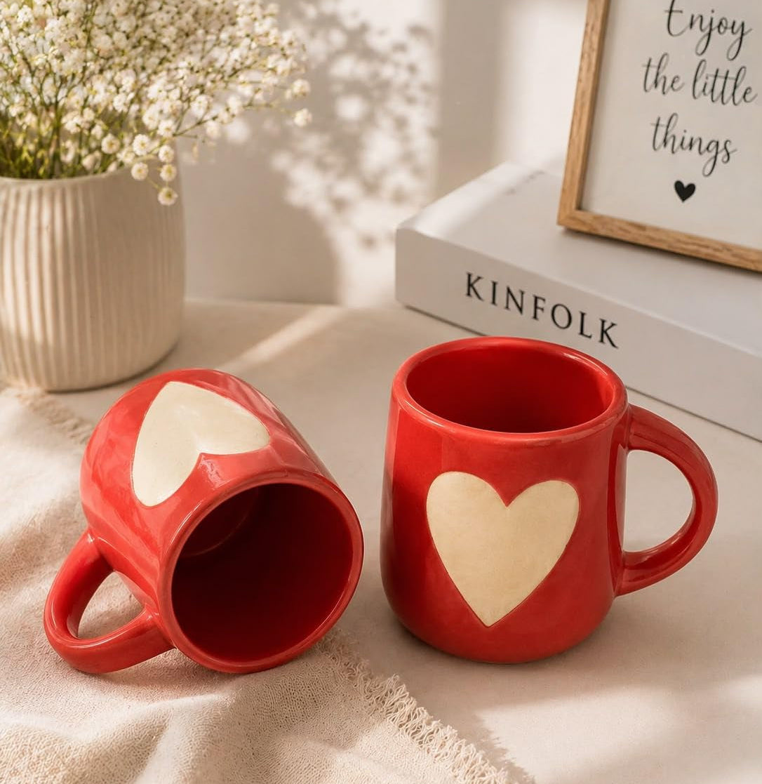 Two red ceramic mugs with cream heart designs, styled on a beige fabric surface with a vase of baby's breath, a book, and a framed sign in the background.