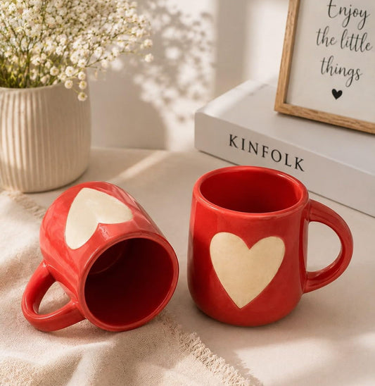 Two red ceramic mugs with cream heart designs, styled on a beige fabric surface with a vase of baby's breath, a book, and a framed sign in the background.