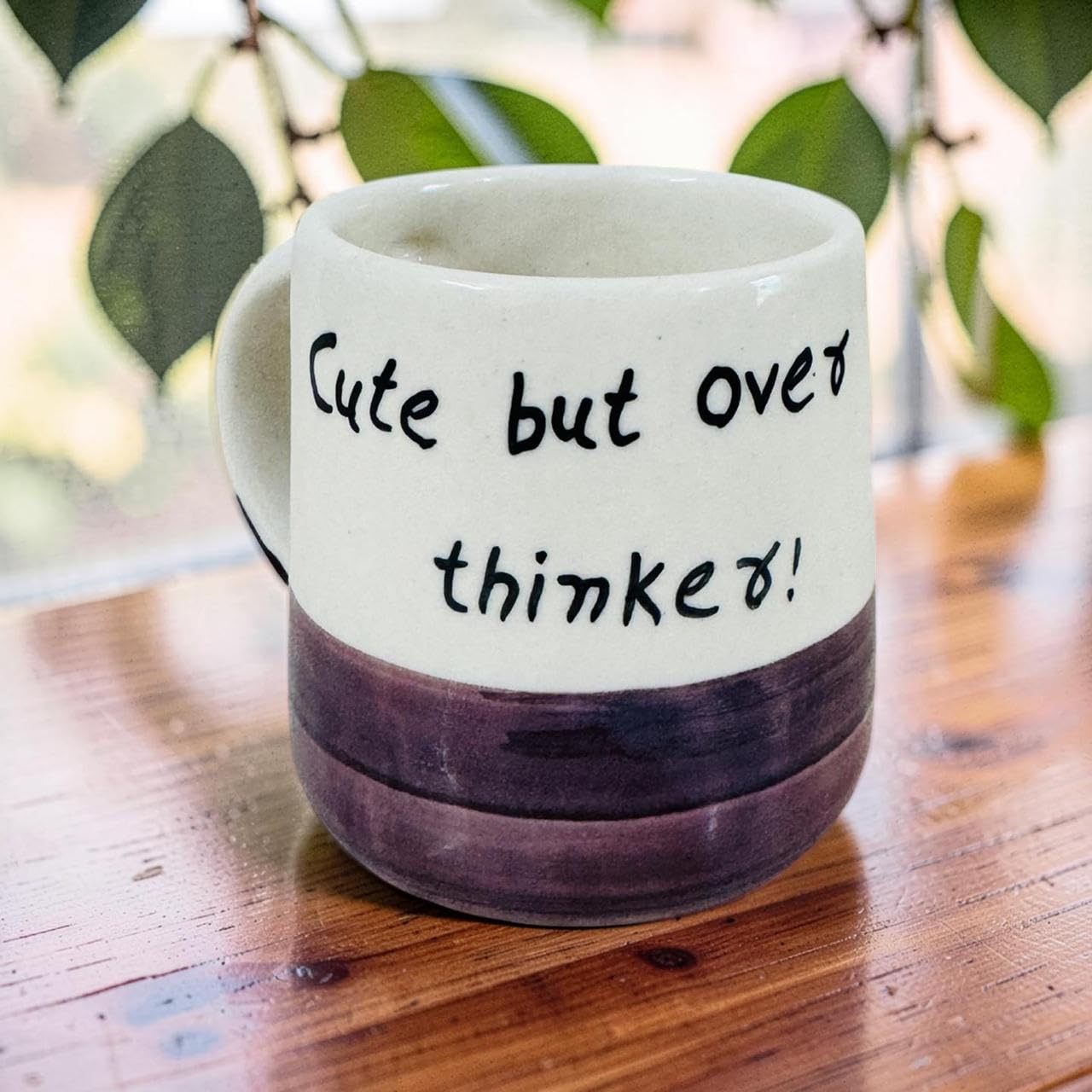 A ceramic mug with a white upper half and dark brown lower half, featuring the text 'Cute but over thought!' in black handwritten-style lettering. The mug is placed on a wooden table with a blurred background of green leaves and bright light.