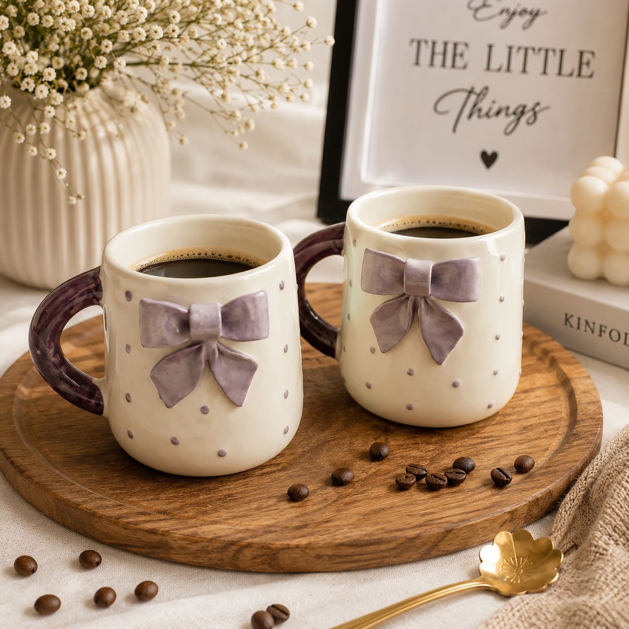 Two ceramic coffee mugs with a cream body, small gray polka dots, and a lavender bow on the front, filled with black coffee and displayed on a round wooden tray with scattered coffee beans.