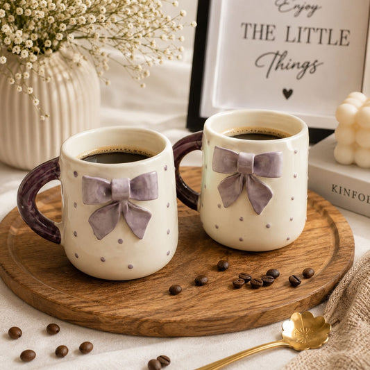 Two ceramic coffee mugs with a cream body, small gray polka dots, and a lavender bow on the front, filled with black coffee and displayed on a round wooden tray with scattered coffee beans.