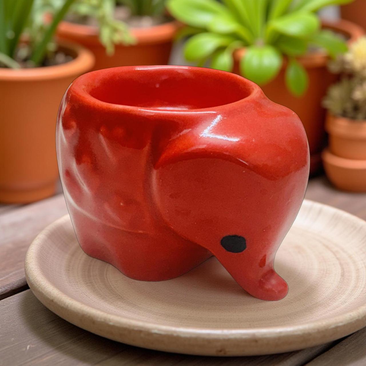 Red elephant-shaped ceramic pot on a beige saucer with plants in the background