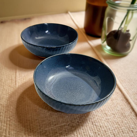 Two blue ceramic bowls on a textured surface with a jar and sticks in the background.