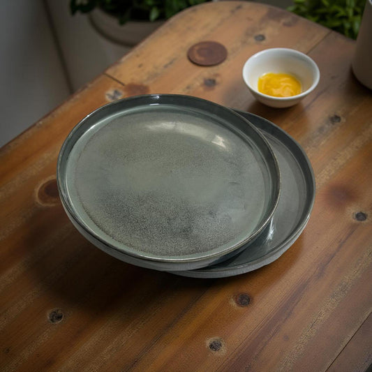 Stack of gray ceramic plates on a wooden surface with a small bowl of yellow liquid in the background.