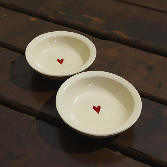 Set of two cream ceramic bowls with red heart in center, placed on dark wooden surface