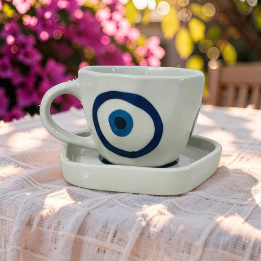 Ceramic cup with blue eye design on a saucer outdoors with flowers in the background