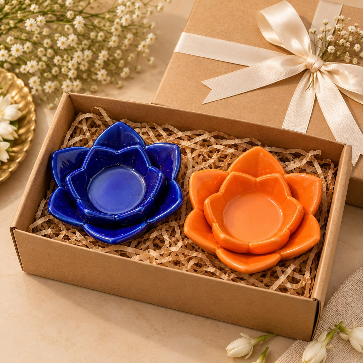 Two ceramic lotus-shaped shallow bowls, one cobalt blue and one terracotta orange, presented in a brown gift box with shredded paper filler. A matching brown gift box with a cream ribbon is visible in the background, along with small white flowers.