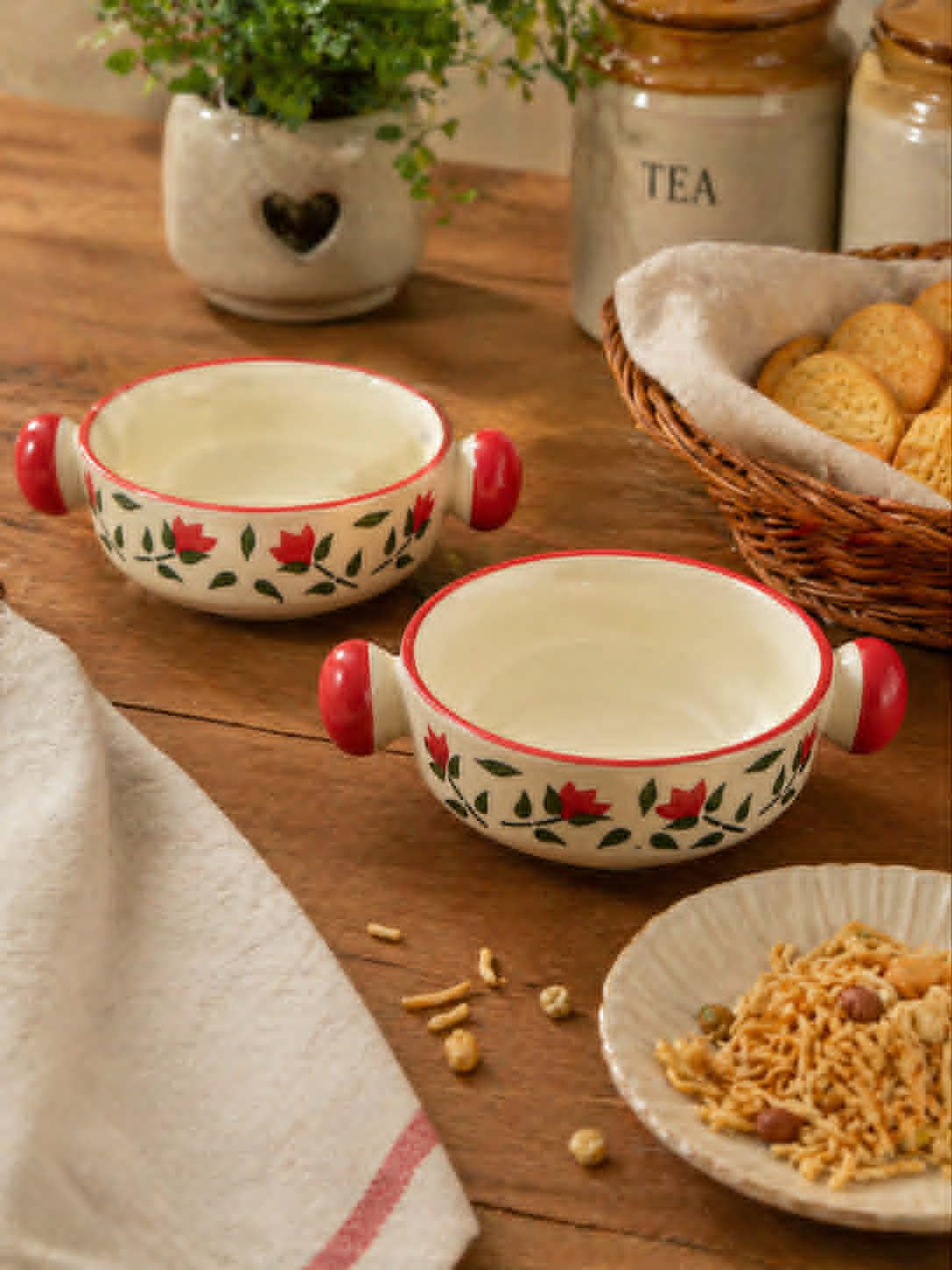 Two cream-colored ceramic bowls with red handles and a red floral pattern, set on a rustic wooden table with a plate of snacks, a basket of cookies, and tea jars in the background.