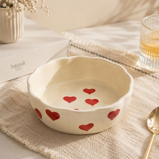 A cream-colored ceramic bowl with a scalloped rim and red heart patterns, styled on a textured beige cloth with a gold spoon, a glass of amber liquid, and a white book in the background.