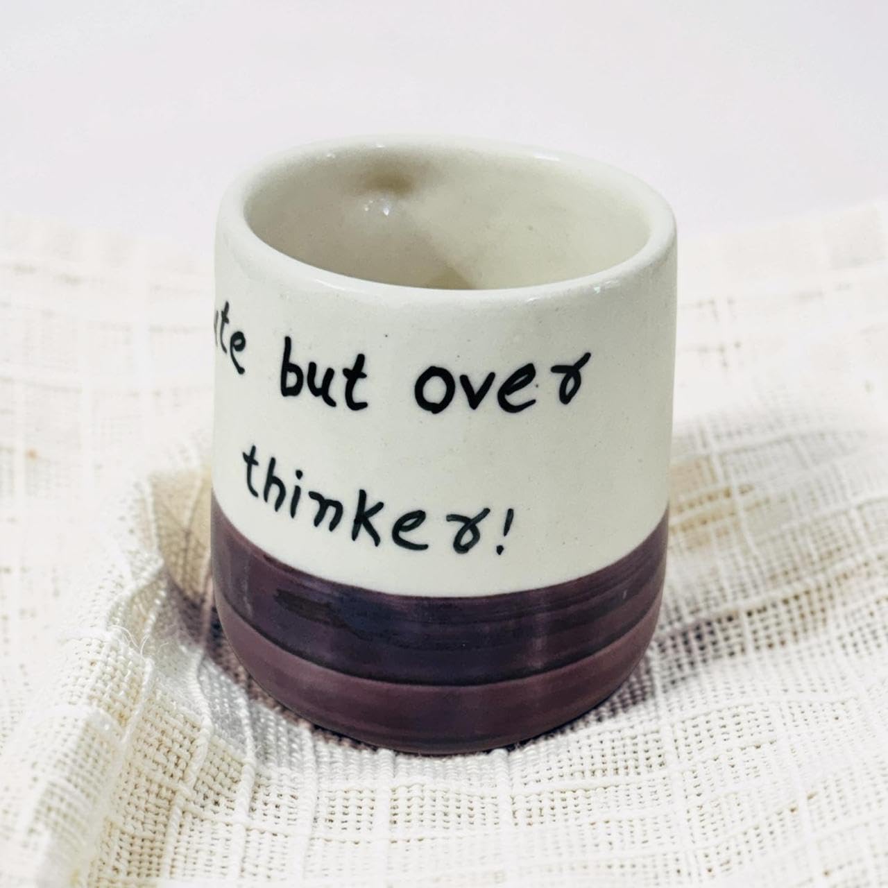 A handmade ceramic cup with a cream-colored upper half, black handwritten text reading 'eat but over think about!', and a dark brown ribbed base, resting on a textured white cloth.