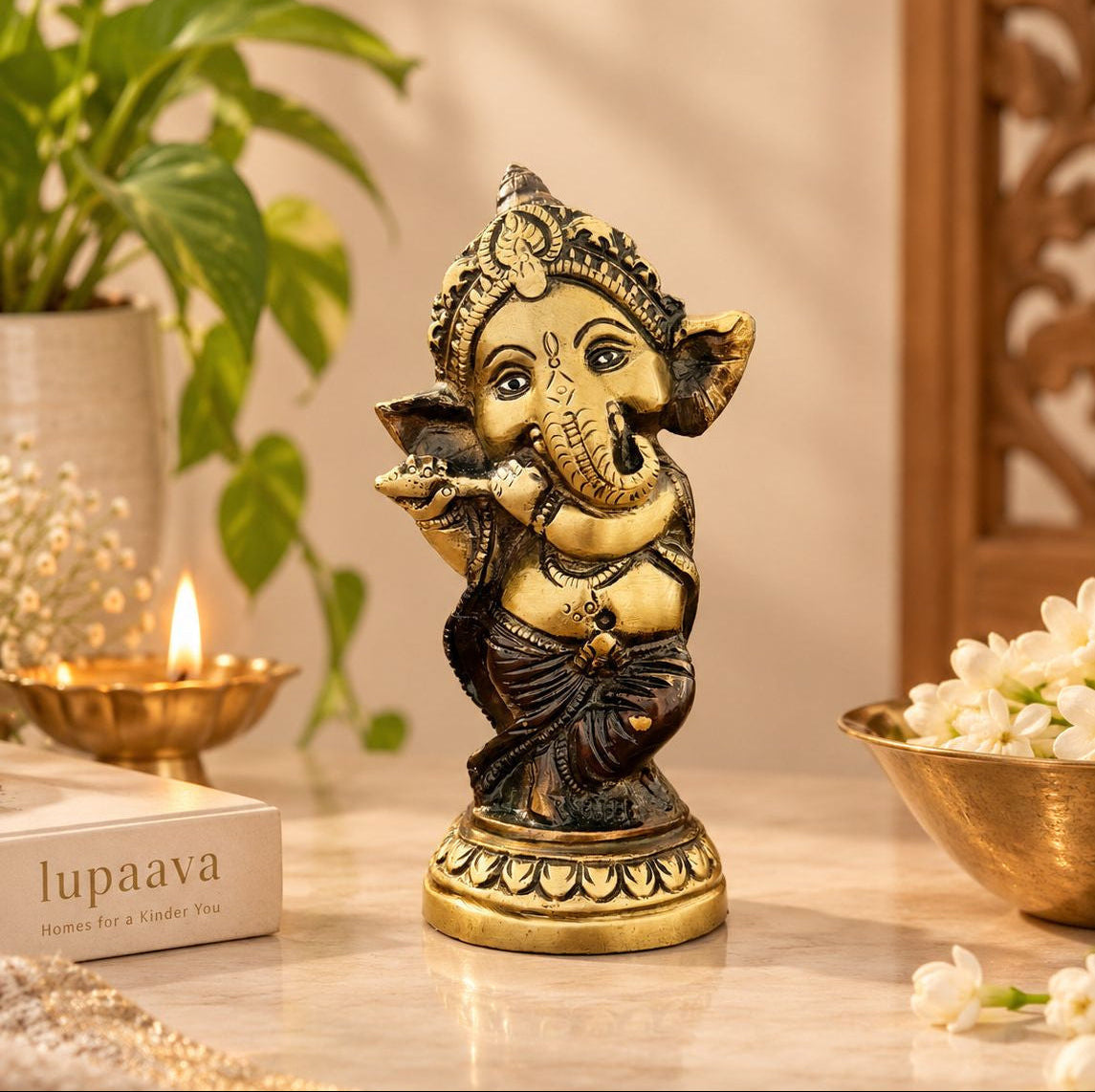 A small brass and black Ganesha statue standing on a marble surface, styled with a lit candle, a book labeled 'lupaava', and white flowers in a warm indoor setting.