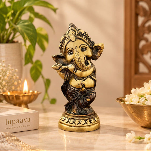 A small brass and black Ganesha statue standing on a marble surface, styled with a lit candle, a book labeled 'lupaava', and white flowers in a warm indoor setting.