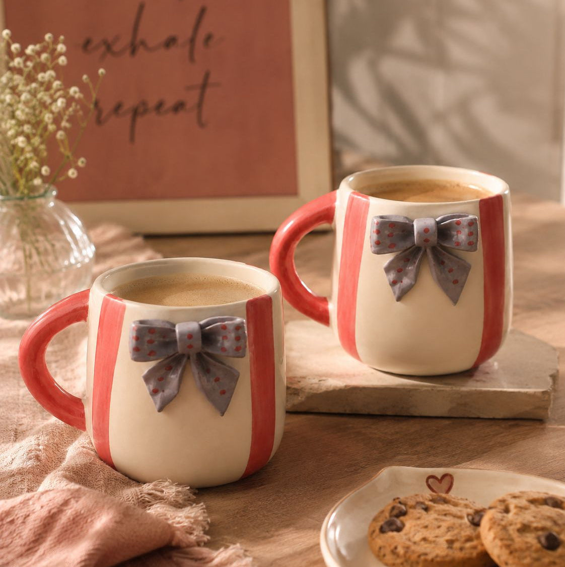 Two ceramic mugs filled with coffee on a wooden table, decorated with coral red and cream vertical stripes and a gray patterned bow. A plate of cookies, a pink sign, and a vase of baby's breath are visible in the background.