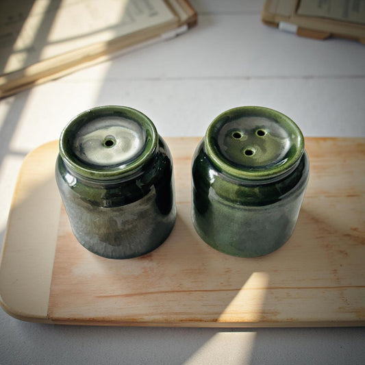 Two green ceramic jars on a wooden surface with books in the background.