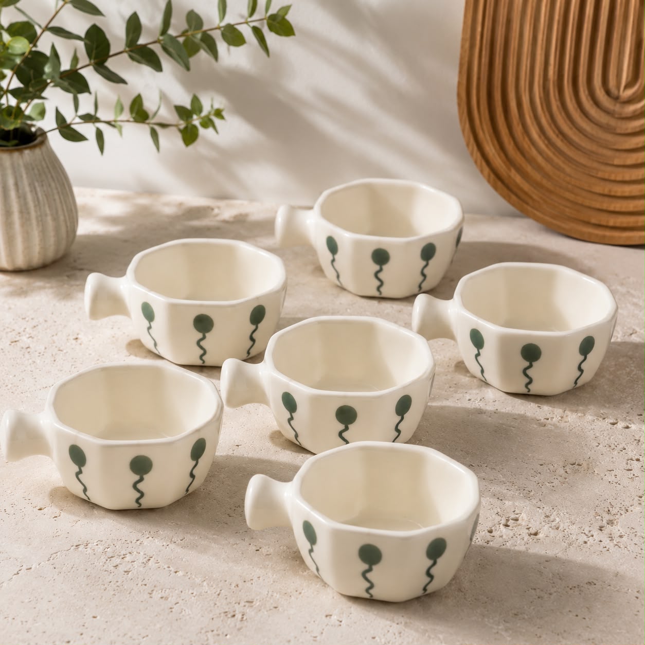 Six small white ceramic cups with handles, decorated with dark green wavy line and dot motifs, arranged on a light stone surface with a vase of greenery and a wooden tray in the background.