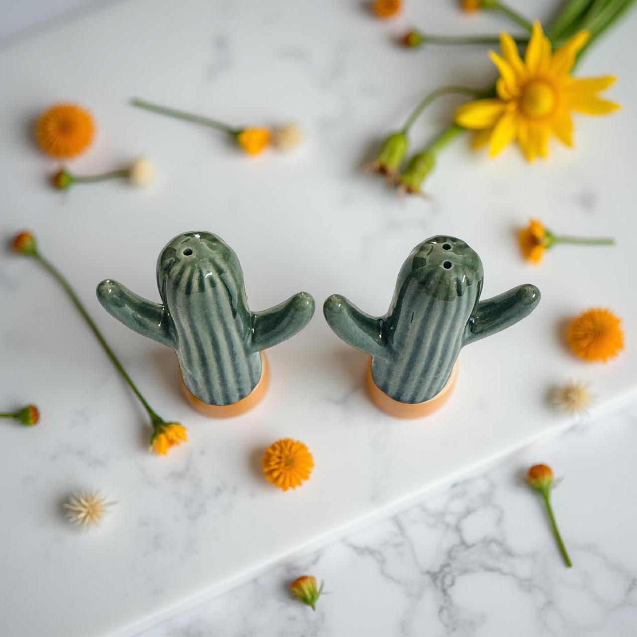 Two cactus-shaped salt and pepper shakers on a marble surface with flowers around.