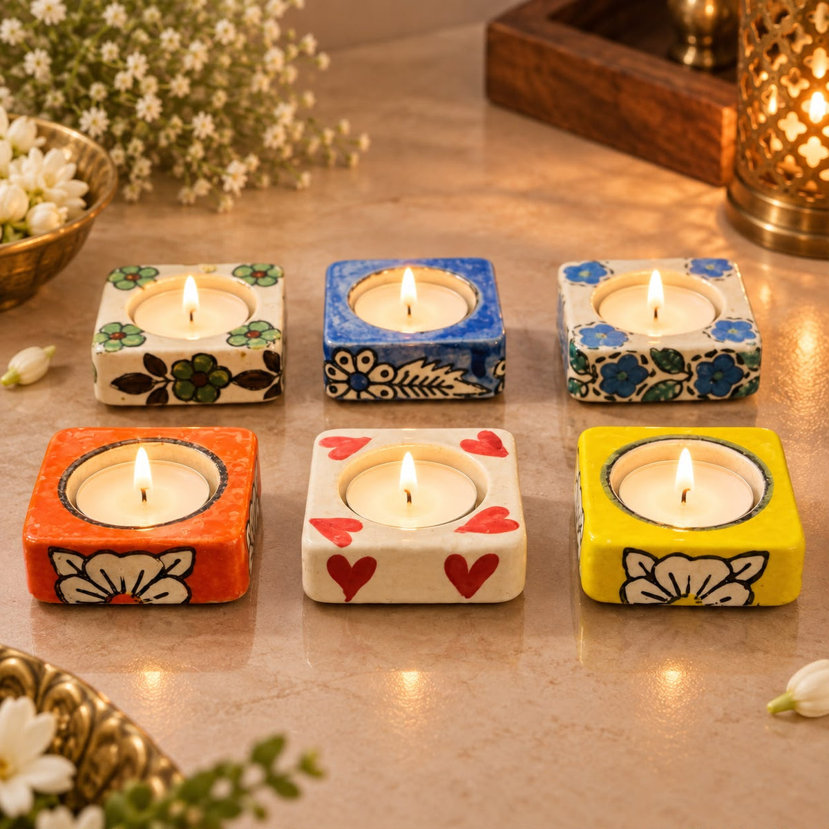 Six square ceramic tealight candle holders with colorful floral and heart patterns, arranged on a tabletop with lit candles inside. The holders feature orange, yellow, blue, and white designs with black painted details.