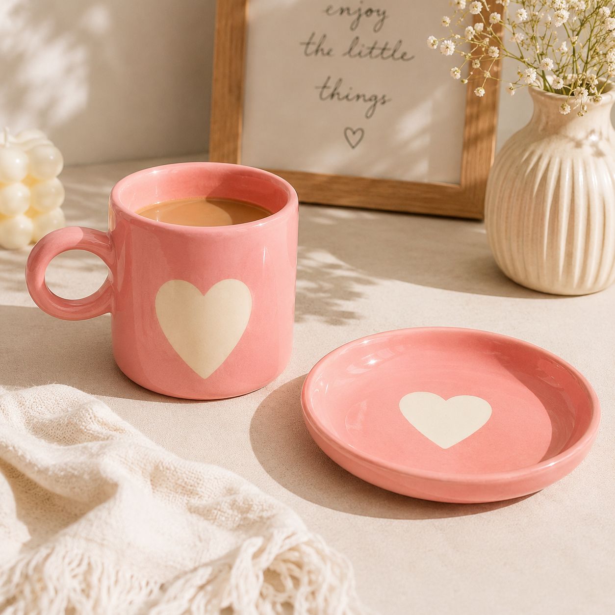 A pink ceramic mug with a matching saucer, both featuring a cream-colored heart design, styled on a beige tabletop with a knit blanket, a small vase of flowers, and a framed print in the background.