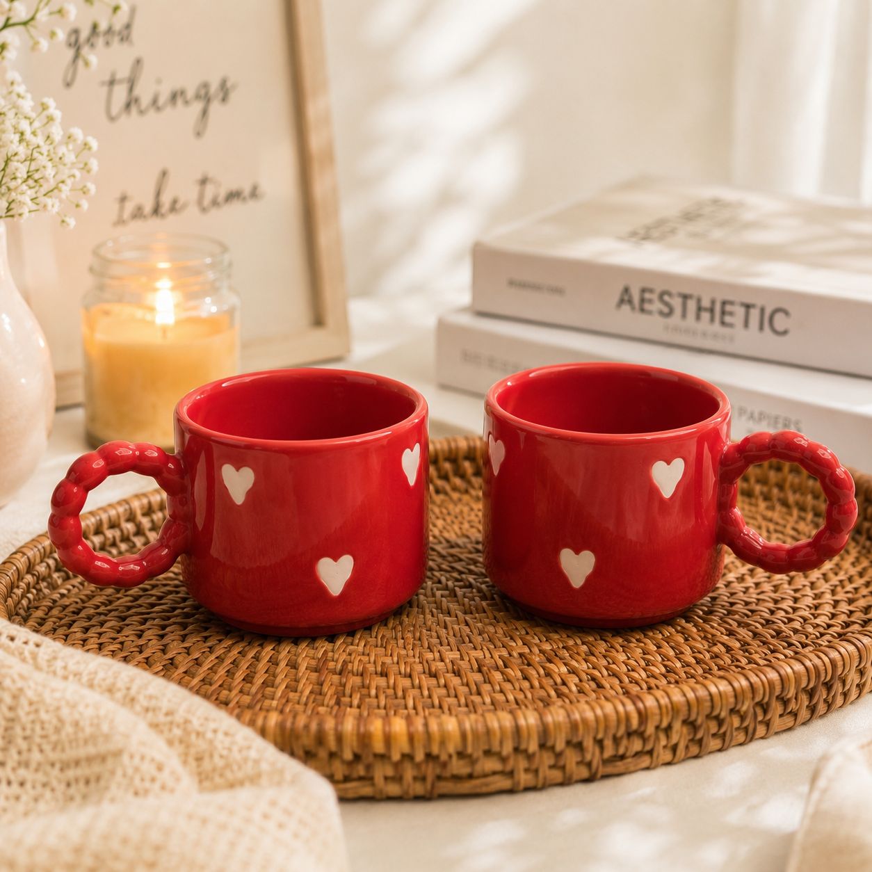 Two red ceramic mugs with textured handles and white heart details, placed on a woven tray in a softly styled setting with books, a candle, and a framed sign in the background.