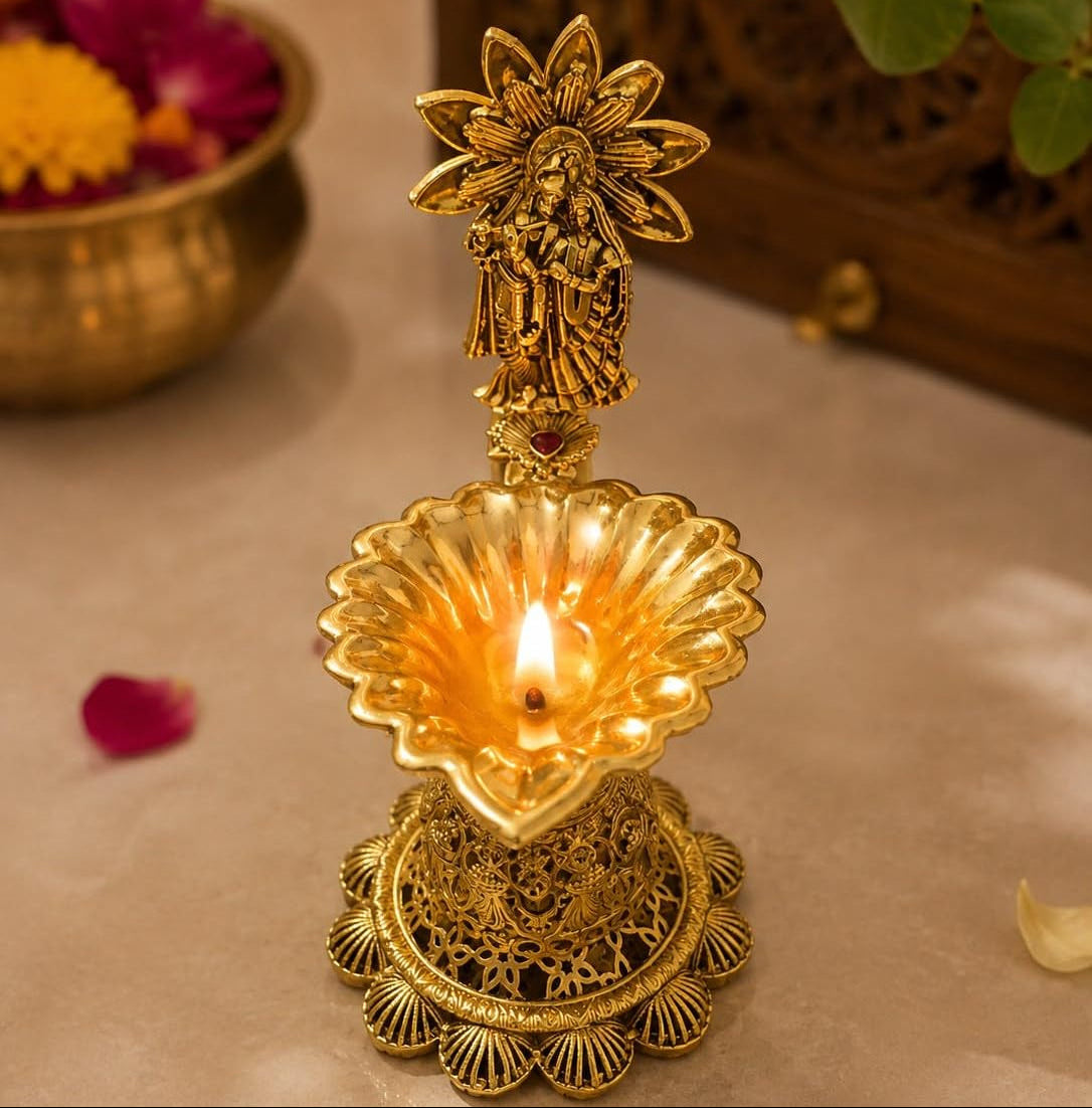 A brass oil lamp (diya) with a decorative figurine on top, lit with a warm flame, set against a softly blurred background with floral elements.