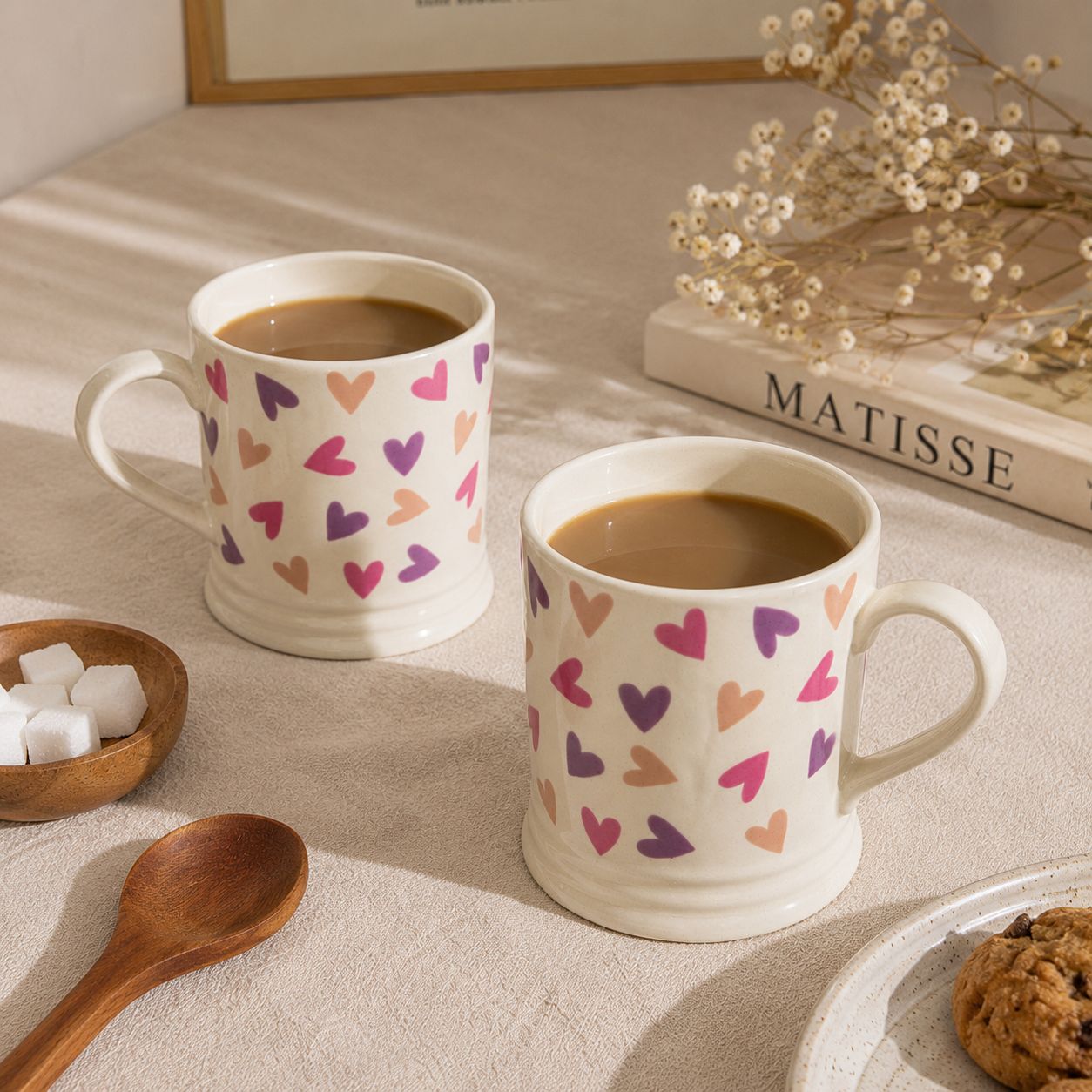 Two mugs with heart patterns filled with coffee on a table with a book and cookies.