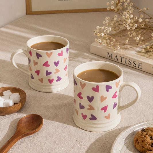 Two mugs with heart patterns filled with coffee on a table with a book and cookies.