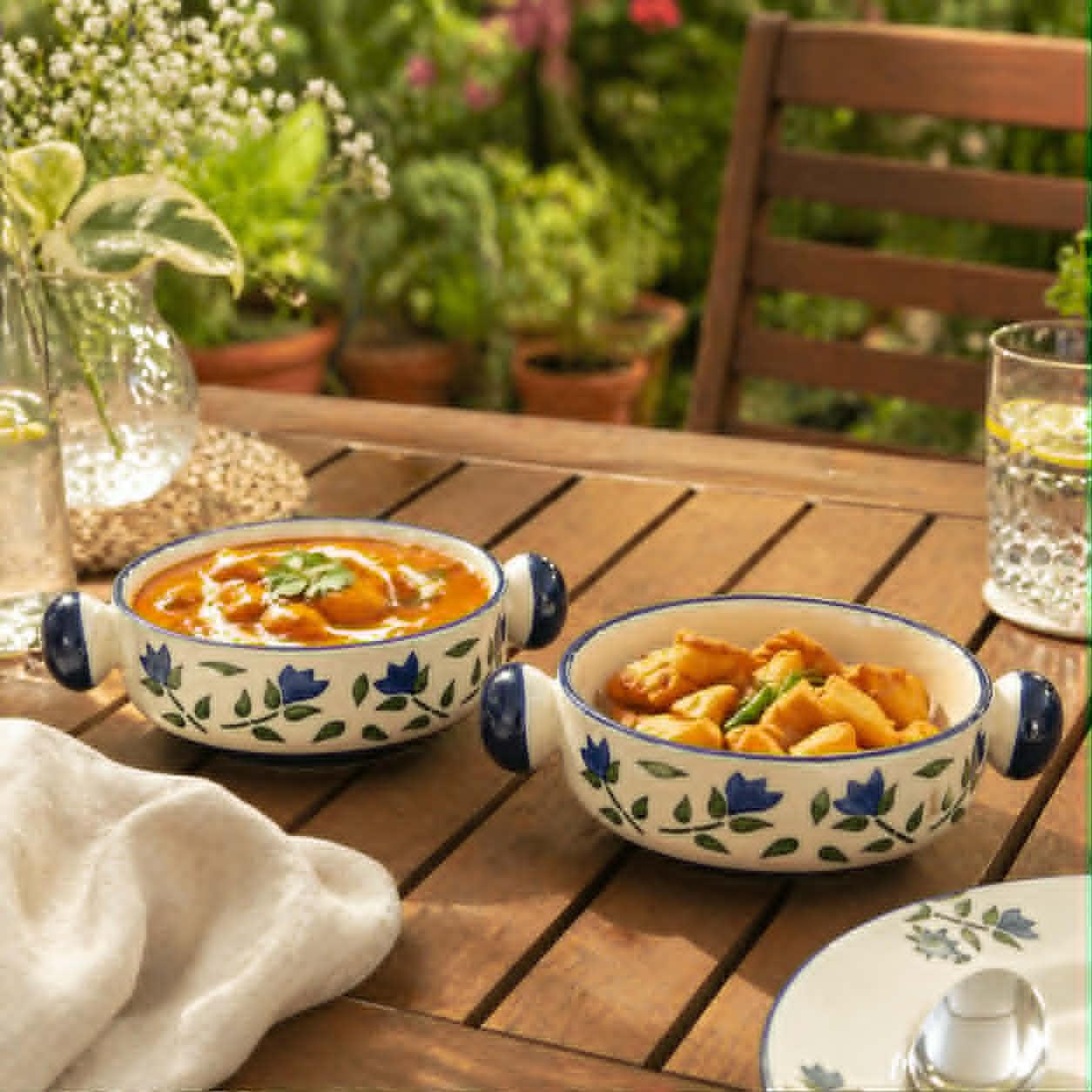 Two ceramic bowls with blue floral patterns and side handles, filled with food, set on a wooden outdoor table with a blurred garden background.