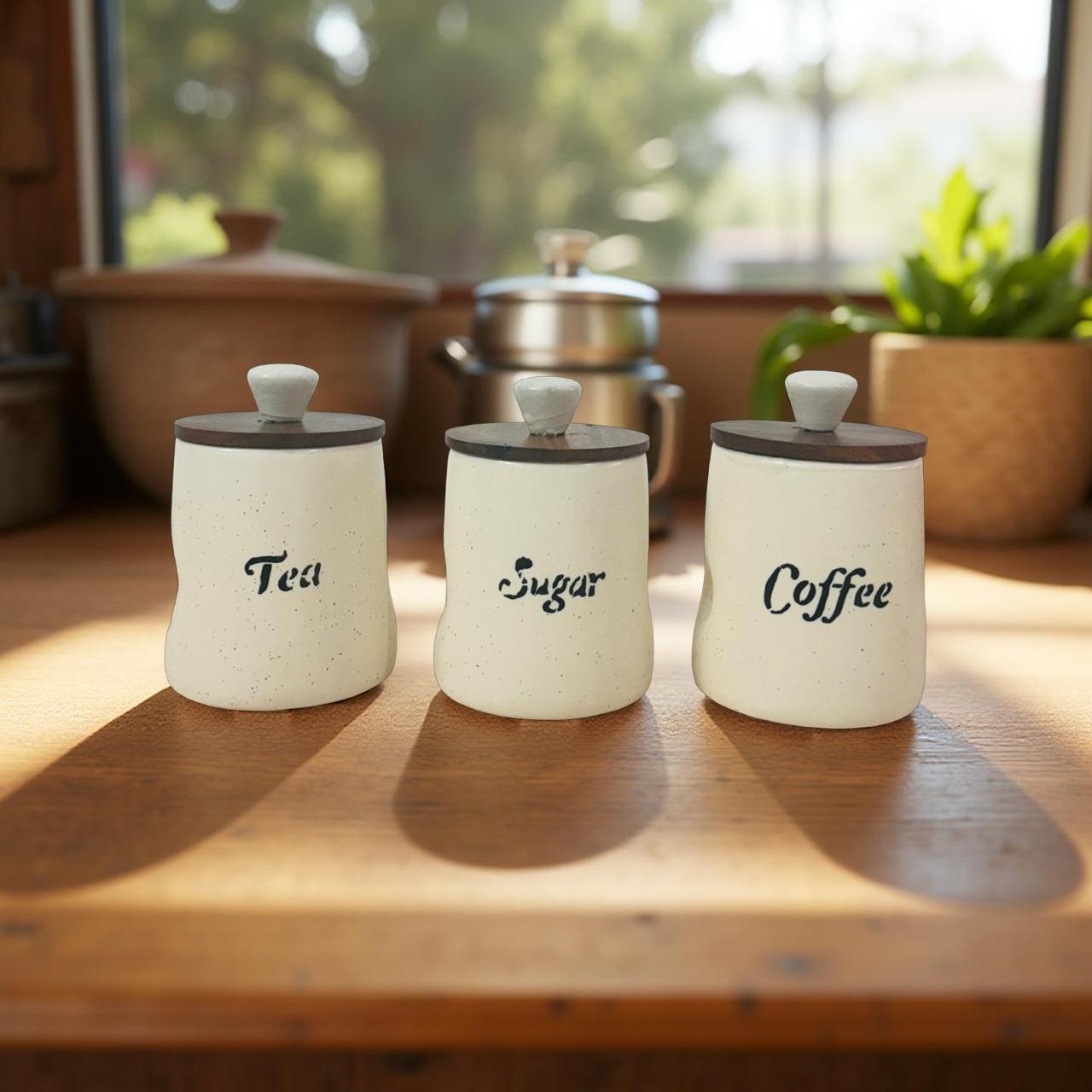Three ceramic canisters labeled 'Tea', 'Sugar', and 'Coffee' on a wooden surface with a blurred natural background.