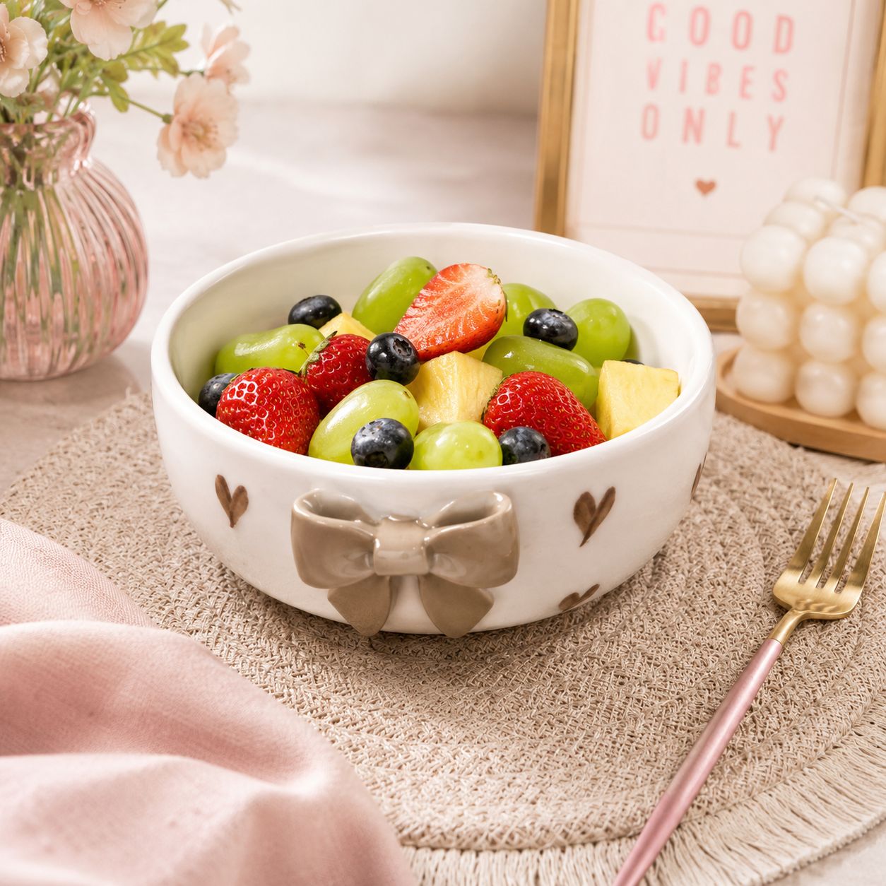 A white ceramic bowl decorated with a beige bow and small brown hearts, filled with fresh fruit including strawberries, grapes, blueberries, and pineapple, styled on a beige placemat with a pink fork and decorative background elements.