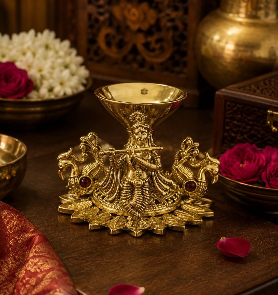 A decorative brass oil lamp holder featuring a detailed Hindu deity figure with two peacock-like creatures on either side and small red gemstone accents, displayed on a dark wooden surface with flowers and ornate background decor.