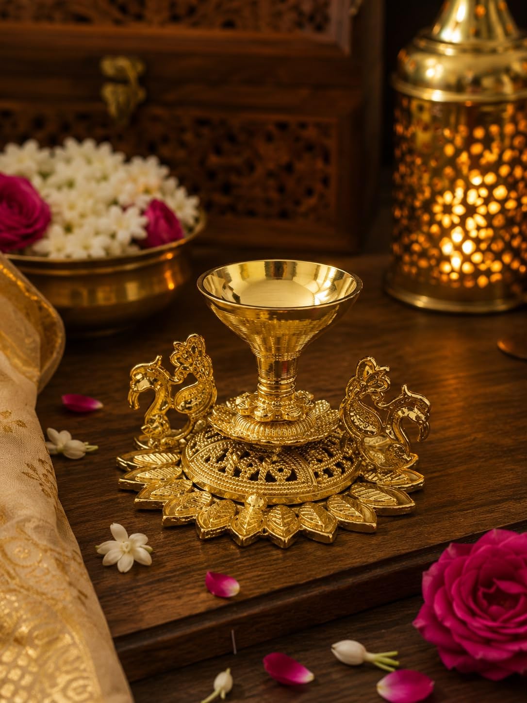 An ornate gold brass oil lamp holder (diya stand) with horse figures on the sides, displayed on a dark wooden surface with decorative flowers and a glowing brass lamp in the background.